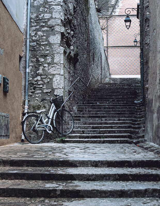 Ruelle avec des vollées de marches basses encadrées par des mures de vieilles pierres, un vélo blanc est attaché sur la main courante à gauche.