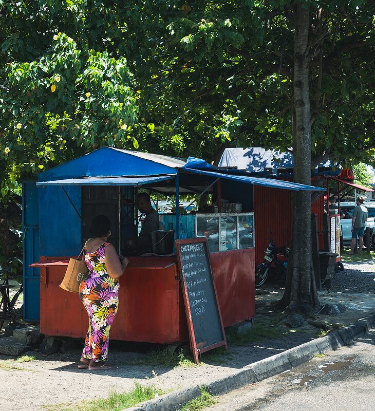 Jeune Mauricienne dans une robe colorée, achetant à manger dans une échoppe à Grand Baie, 2026.